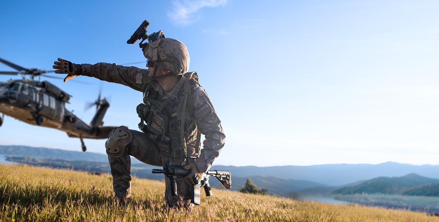 Military man dressed in a combat uniform, kneeling on ground holding a weapon and signaling to a helicopter 